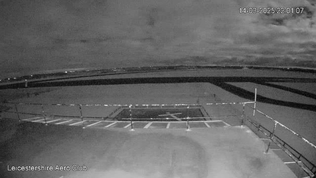 A dark night scene showing the Leicestershire Aero Club area. In the foreground, there is a helipad with a white cross marking its center. Surrounding the helipad are several white parking lines. Beyond the helipad, a flat expanse extends toward the horizon, with faint lights from the distance suggesting nearby buildings or structures. The sky is largely cloudy, contributing to the overall dark ambiance of the scene. The timestamp in the top right corner indicates the image was captured on March 14, 2025, at 10:01 PM.