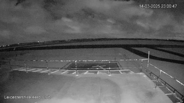 A dark, nighttime image from a webcam showing an airport runway area. The foreground features a fenced-off section with a runway marking and white lines, indicating landing or takeoff areas. In the background, the sky is partly cloudy with some distant lights visible, suggesting nearby buildings or roads. The date and time display is located in the upper right corner of the image.