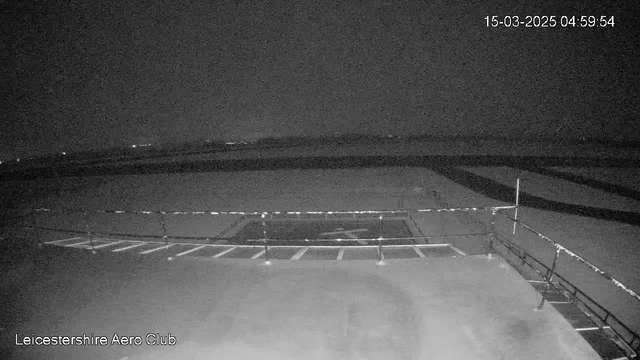 A low-light webcam image shows a view of an airfield at dawn. The scene is predominantly dark with faint outlines of the runway visible. A parked helicopter pad with a marked "H" is in the foreground, and there is a railing along the edge of the platform. The background is hazy with distant lights that suggest the presence of buildings or vehicles. The date and time are displayed in the top right corner: 15 March 2025, 04:59:54.