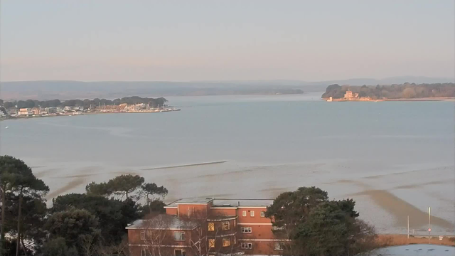 A tranquil view of a bay with calm water reflecting soft morning light. In the foreground, a low-rise brick building with several windows is partially obscured by trees. Across the bay, a line of small boats is docked along a coastal area that has some buildings and greenery. In the distance, a large structure resembling a castle or historic building is visible on a landmass surrounded by water, with rolling hills beyond. The sky is clear and light, indicating an early morning or late afternoon scene.