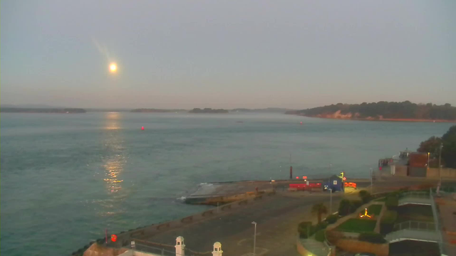 A tranquil coastal scene at dusk featuring calm waters reflecting soft light from the rising moon. In the foreground, a concrete path runs along the shore, bordered by green landscaping. A dock area is visible with boats and a blue building. To the right, there are hints of nearby structures and trees lining the distant shoreline. A few small red buoys are scattered across the water, enhancing the peaceful atmosphere.