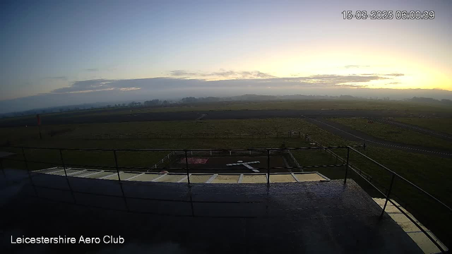 A wide view of an airfield taken from a webcam at the Leicestershire Aero Club during early morning. The foreground features a railing and part of a flat surface. Below the railing, an 'X' marking is visible on the ground, indicating a landing area. In the background, the grassy airfield stretches out, with scattered patches of dirt and several indistinct shapes, likely vehicles or markers, against a cloudy sky that transitions from shades of blue to yellowish light as the sun rises. The date and time displayed in the corner indicate it is March 15, 2025, at 6:00 AM.