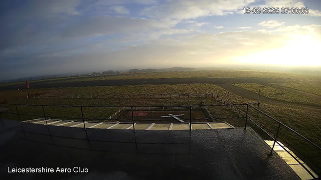 A view from a webcam at Leicestershire Aero Club shows an expansive landscape during sunrise. The foreground includes a portion of a concrete balcony with a metal railing. In the distance, an open field stretches out toward a runway, which is visible as a strip of gray asphalt. To the left, there is a red windsock indicating wind direction. The sky above features soft clouds illuminated by the rising sun, casting a warm glow over the scene.