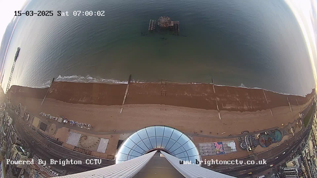 A view from a high vantage point facing the sea. Below, there is a wide sandy beach with gentle waves lapping at the shore. In the water, a partially submerged pier is visible, while further along the beach, there are various structures, including amusement park rides and other buildings. The sky is lightening with the dawn, and the image is time-stamped with the date and time at the top. The bottom features a curved glass structure, likely part of a building.