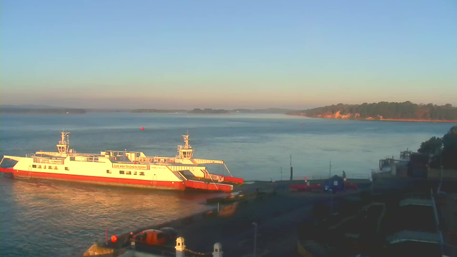A white and red ferry is docked at a waterfront, with calm waters reflecting the morning light. In the background, a shoreline with trees and cliffs stretches along the horizon under a clear blue sky. Various objects, including boats and equipment, are visible along the dock.