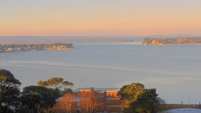 A serene waterfront scene at sunrise, featuring calm waters reflecting soft pastel hues. In the foreground, there are hints of trees and a building with an orange facade. To the left, a harbor with boats is visible, while the right side reveals distant land and a building on an island. The sky is gradually lightening, creating a tranquil atmosphere over the landscape.