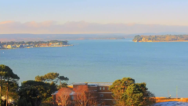 A tranquil waterfront scene depicting a body of water under a clear sky. On the left, a small coastal town with colorful buildings and boats docked along the shore. In the foreground, trees and shrubs add greenery, while a few buildings are partially visible. In the background, gentle hills and islands can be seen faintly in the distance, creating a serene landscape.