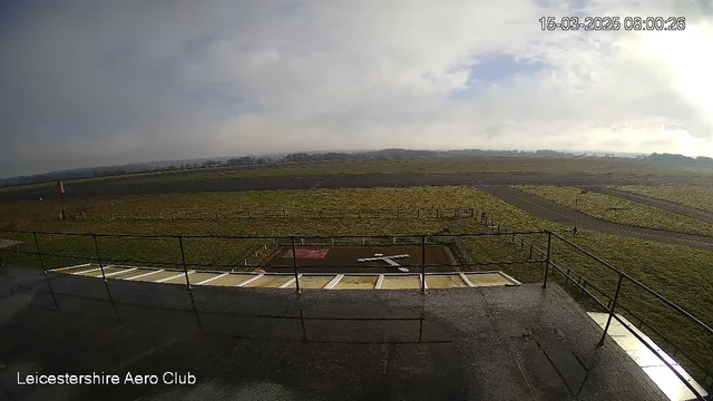 A view from a webcam overlooking a grassy airstrip. In the foreground, there is a helipad marked with a white cross layout on a reddish dirt area. Surrounding the helipad is a grassy field with white fencing. In the background, the landscape is mostly flat, with a few distant hills under a cloudy sky. The time stamp shows the date and time as March 15, 2025, at 08:00:26.