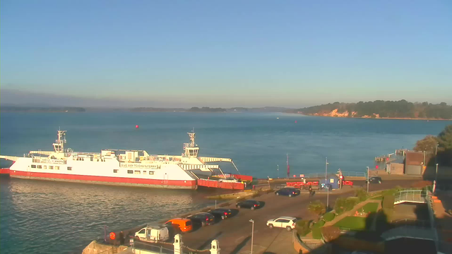 A white and red ferry is docked at a harbor with clear blue water. The background features a shoreline with green trees and cliffs. Various vehicles are parked along a road near the water, and there are traffic signs and barriers visible. The sky is bright and clear, indicating a sunny day.