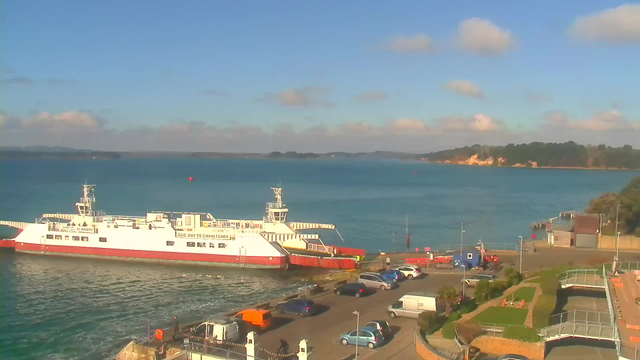 A ferry is docked at a terminal with a vibrant blue sea in the background. The sky is mostly clear with a few fluffy clouds. A parking area with various cars, including an orange van, is situated in the foreground. To the right, there’s green landscaping with pathways and steps leading towards the terminal. In the distance, a wooded shoreline is visible along the horizon.