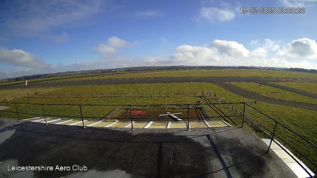 A view from a rooftop at Leicestershire Aero Club showing a grassy area with a cross-shaped arrangement made of white materials on the ground. Surrounding the area are wooden fences and a tarmac runway extending toward the horizon. The sky is partly cloudy, with patches of blue visible. The scene is illuminated by daylight.