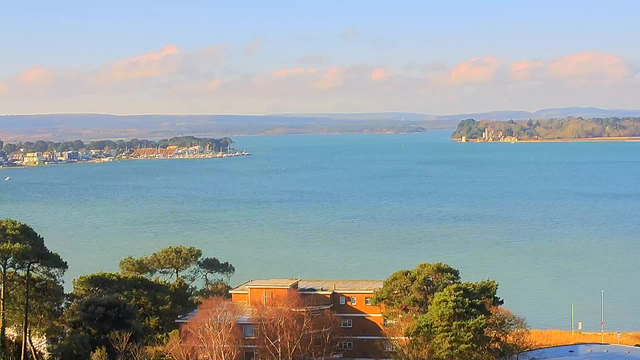 A tranquil view of a body of water on a clear day. The water appears calm with a light blue hue, reflecting the sky. In the distance, a shoreline features several small buildings and trees. There are a few boats visible on the water, and small white markers or buoys are scattered throughout. The landscape is framed by gentle hills in the background, with a mix of greenery and patches of brown land. The foreground shows a low building with a flat roof and some shrubs and trees. A few clouds are present in the sky, suggesting a pleasant day.