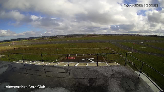 A view from a webcam at the Leicestershire Aero Club. The foreground shows a flat rooftop with a railing on the right side, overlooking a grassy landscape below. In the center, there is a helipad marked with a white cross. The background features a wide, open sky filled with white clouds and patches of blue. The ground below has a mix of grass and tarmac areas, with some blurred outlines of fences and runways in the distance. The timestamp in the corner shows the date and time as 15 March 2025 at 10:00:28.