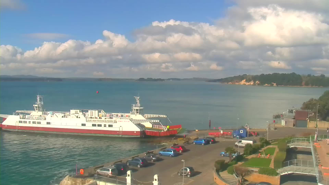 A ferry is docked at a quiet harbor. The ferry is predominantly white with red accents, and a crew member can be seen on the deck. The water is a calm blue, reflecting fluffy white clouds in the sky. In the foreground, there is a parking lot with several cars and some greenery. The shoreline features trees and a sandy beach in the distance, with a gentle slope leading down to the water's edge.