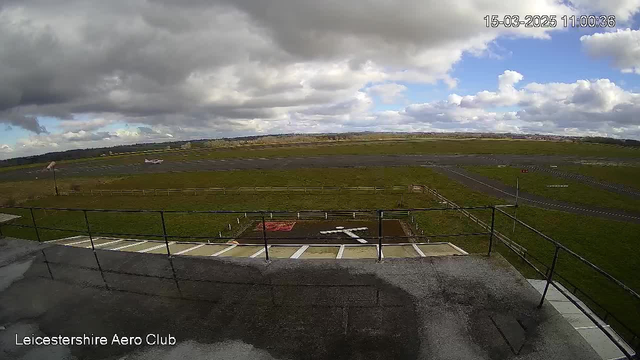 A view from a webcam showing a wide expanse of an airfield under a partly cloudy sky. In the foreground, there is a railing and a patterned surface with a helicopter landing pad marked by white lines on brown ground. The background features grass and a paved runway with a small aircraft visible towards the left. The horizon is lined with distant hills and fields, all under a mix of gray and blue sky.