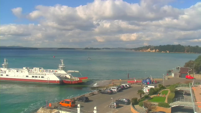 A ferry approaches the shore in a clear blue sea, with gentle waves lapping against the dock. The sky is mostly sunny with fluffy clouds scattered throughout. A parking lot is visible on the right, filled with vehicles, including an orange one. There are some greenery and walkways leading towards the water, and distant land visible on the horizon.