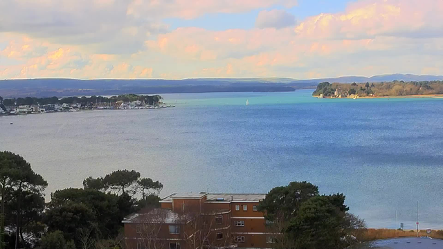 A scenic view of a tranquil body of water under a partly cloudy sky. In the foreground, there are trees and a brown building. To the left, a small marina with white boats is visible along the shoreline. The water exhibits varying shades of blue, leading to a distant green island on the right, where some structures can be seen. The horizon features rolling hills in the background.