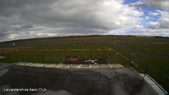 A view from a webcam positioned at the Leicestershire Aero Club. The image shows an expansive grass field divided by runways. The sky is mostly cloudy with patches of blue. In the foreground, there is a concrete area with a white cross marking. A railing runs along the edge of the platform where the camera is located, providing a vantage point over the airstrip. In the distance, the horizon features fields and a few scattered clouds.