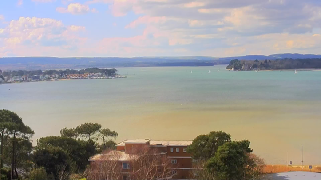 A scenic view of a coastal area featuring calm water that transitions from blue to green, bordered by trees and small buildings. In the distance, a marina with several boats is visible along with a shoreline lined with houses. The sky is partially cloudy with soft, white clouds scattered above, and rolling hills are seen in the background.