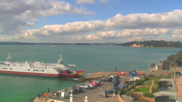 A scenic waterfront view featuring a large ferry docked at a pier, with vehicles parked nearby. The background includes a calm, turquoise body of water with gentle waves and white clouds scattered across a blue sky. In the distance, there are green hills and a sandy coastline. The foreground has a few palm trees and landscaped areas.