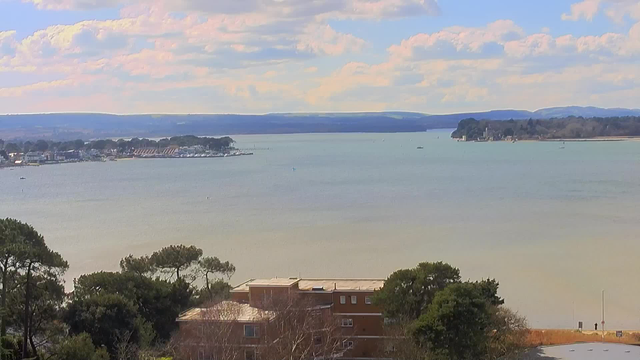 A panoramic view of a calm body of water extends towards the horizon, with gentle ripples reflecting soft, cloudy skies. In the foreground, a building with a flat roof is partially obscured by surrounding trees. Further back, a small marina is visible, lined with boats, and beyond it, a distant landscape of rolling hills fades into the sky. The overall atmosphere is serene and picturesque.