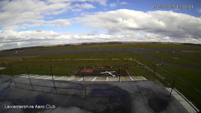 A view from a webcam positioned on a building at an airfield. In the foreground, there is a flat surface with lines and a red and white square marking. Beyond this area, a grassy field stretches towards a runway, which is paved and extends into the distance. The sky is partly cloudy with blue patches visible, and there is a small structure visible at the edge of the airfield.
