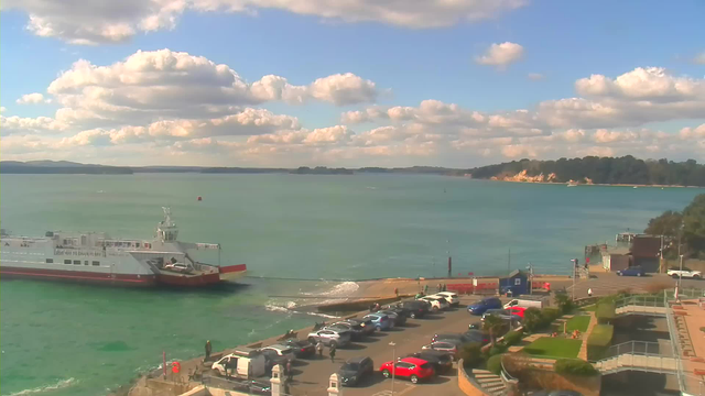 A large ferry is docked at a port with a ramp extending towards the shore. The water is a calm turquoise, reflecting the blue sky filled with fluffy white clouds. In the foreground, there is a parking area with various cars, including red and white vehicles. There are a few people walking near the water's edge and a small boat is visible in the distance. To the right, lush greenery and steps leading down are seen, along with a few structures, including a building near the edge of the water.