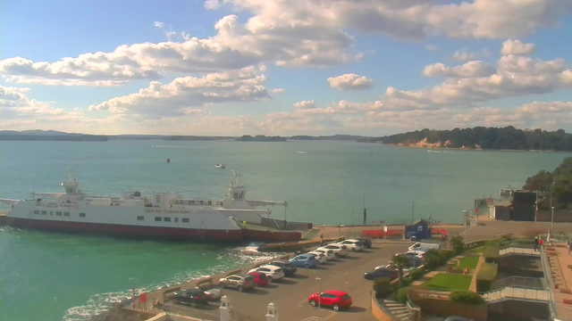 A scenic view of a harbor with a ferry docked on the left side. The ferry is white with red accents and has the words "SAVE WAY TO CHAIN FERRY" visible. The water is calm and turquoise, with a few small boats in the distance. In the foreground, there is a parking area with several cars, mostly in shades of blue, red, and white. Lush greenery is visible on the right side of the image, along with steps leading down to the water. The sky is bright with fluffy white clouds scattered across a blue backdrop.