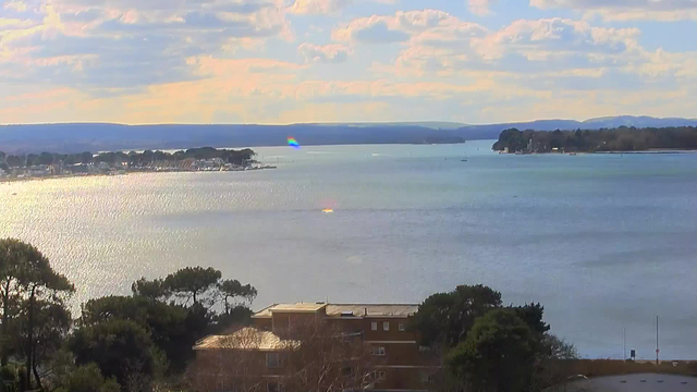 A serene view of a body of water under a partly cloudy sky. The water reflects various shades of blue and silver, with gentle ripples and a few small boats visible in the distance. On the left side, a coastal area features buildings and trees. In the background, mountains line the horizon. The scene is tranquil and picturesque, evoking a sense of calm.