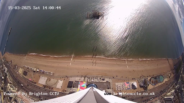 Aerial view of a beach and seaside area on a sunny day. The sand stretches along the bottom of the image, with a few small figures walking in the distance. A gentle wave can be seen lapping at the shore. In the water, a pier extends outwards, with a structure visible at its end. The coastline includes amusement rides and colorful beach umbrellas, showcasing a bustling beachfront atmosphere. The sea is calm and reflects sunlight, creating a shimmering effect. The date and time are displayed in the top left corner.