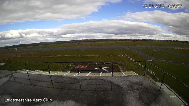 A view from a webcam at the Leicestershire Aero Club, capturing a large grassy area under a partially cloudy sky. In the foreground, there is a railing along a terrace, with a marked landing area featuring a white cross shape and a red square. A paved runway is visible in the background, along with several signs and a nearby fence separating the landing area from the field. The scene is bright, with varying cloud formations overhead.
