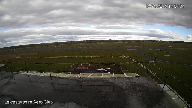 A view from a webcam showing a landscape with a large open field. The foreground features a paved area with a large white cross marking, indicating a landing zone. To the left, there is a railing, and beyond that, a grassy field stretches out, bordered by a white fence. In the background, a portion of the asphalt runway is visible, along with several clouds in a mostly overcast sky. The scene is tranquil, with a hint of industrial presence from the airport facilities.