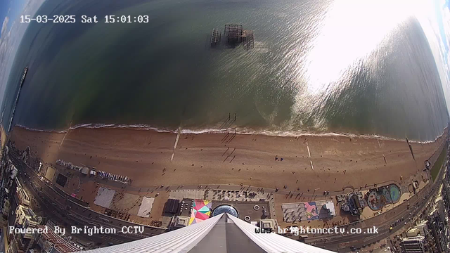A view from a high vantage point overlooking a sandy beach beside a blue sea. Several people are walking along the shore, with some larger structures, including a pier with a partially collapsed building, visible in the water. Below the beach, there are various attractions and activities, including colorful tents and a circular pool. The sky is partially cloudy, with sunlight reflecting off the water surface. The image includes a timestamp and a watermark for Brighton CCTV.