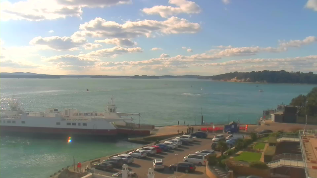 A sunny coastal scene featuring a harbor. In the foreground, a large white ferry is docked next to a small pier, surrounded by a few people. Vehicles are parked in a nearby lot, and some greenery is seen in the landscape. The water appears calm and blue, reflecting the sunlight, with a backdrop of distant hills and a partly cloudy sky.
