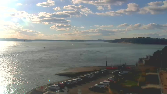 A scenic view of a bay under partly cloudy skies. The water is shimmering in the sunlight, with gentle waves. In the foreground, there is a parking area filled with parked cars. A dock extends into the water, and some people can be seen walking on it. In the distance, hills rise along the shoreline, creating a tranquil atmosphere.