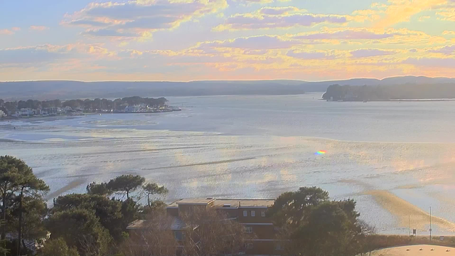 A scenic view of a calm body of water reflecting soft colors from the sky at sunset. The scene features a mix of blues and light oranges, with wispy clouds scattered throughout. In the foreground, there are trees with visible foliage, and in the distance, a small shoreline with several boats and buildings can be seen along the water's edge, fading into the horizon where rolling hills meet the sky.