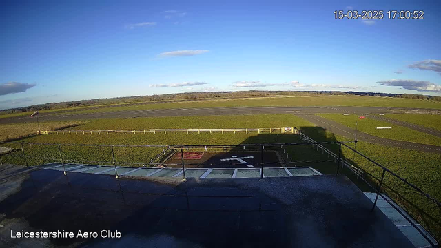 A wide view of an airfield under a clear blue sky. The foreground features a runway and a helicopter landing area with a painted cross. There is a fence surrounding part of the airfield, and the horizon shows green fields and scattered clouds. A flag on a pole gently flutters in the breeze.