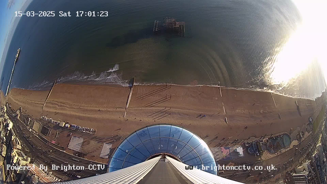 A panoramic view from a high vantage point overlooking a beach. The scene captures the shoreline with golden sand and gentle waves lapping at the edge of the water. A pier extends into the sea, and in the distance, a partially submerged structure is visible. To the left, a section of the boardwalk and several buildings are in view, showcasing the seaside's recreational area. The sky is clear with some clouds, and sunlight glimmers on the water's surface, creating a bright reflection.
