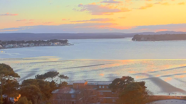 A scenic view of a calm body of water during sunset, with soft pastel colors of orange, pink, and purple reflecting on the surface. In the foreground, there are some buildings and trees. The horizon is lined with distant hills and a few boats can be seen in the water near a small harbor, while the sky features scattered clouds.