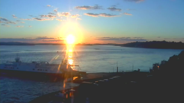 A scenic view of a waterfront during sunset. The sun is setting on the horizon, casting a warm golden light across the water, which reflects shimmering rays. To the left, a large ferry boat is docked at the pier, surrounded by gentle waves. A shoreline with trees and hills is visible in the background, and scattered clouds add texture to the vibrant sky. The foreground has a dark outline of parked vehicles along the waterfront.