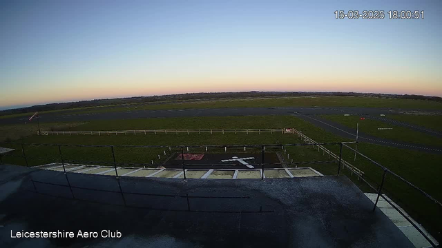A view from a height overlooking an airfield at dusk. The sky transitions from light blue to orange near the horizon. In the foreground, a railing surrounds a flat surface. There is a symbol on the ground resembling a helicopter pad, and some markings on the tarmac. Beyond, the airfield has several grassy areas and a few white fences delineating the boundaries, with a windsock visible on the left.