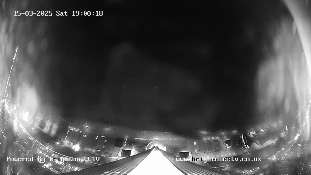 A black and white image shows a high-angle view from a webcam overlooking a city street at night. The scene is largely dark, with faint outlines of buildings and structures illuminated by streetlights. In the lower portion of the image, a long, narrow building or structure is visible, tapering towards a point. The date and time in the top left corner indicate it is March 15, 2025, at 19:00:18. The bottom left corner displays the text "Powered By Brighton CCTV," and the bottom right corner shows the website "www.brightoncctv.co.uk."