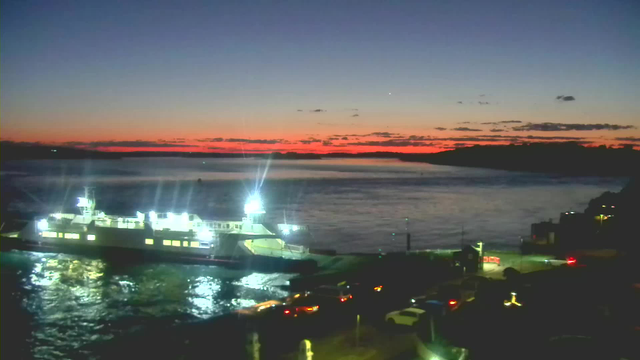 A tranquil scene at dusk featuring a ferry docked at a waterfront. The ferry is illuminated with bright lights, reflecting on the water's surface. The sky transitions from deep blue to vibrant orange along the horizon, with scattered clouds. In the foreground, vehicles are parked near the shoreline, while a few structures line the dock, creating a peaceful evening atmosphere.