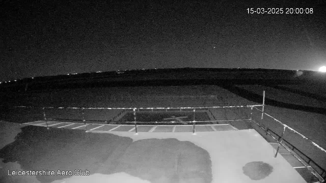 A dark scene captured by a webcam at night, showing a runway area with a faint outline of multiple aircraft taxi lines on the ground. There is a railing in the foreground and a paved surface that appears slightly wet. The sky is mostly dark with a few distant lights visible on the horizon, and the time and date are displayed at the top right corner, indicating March 15, 2025, at 8:00 PM.