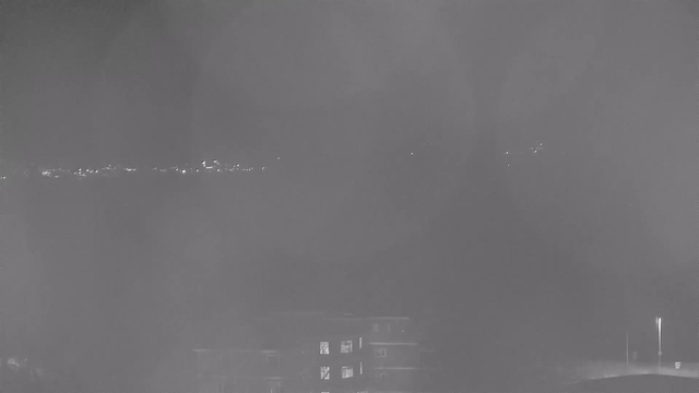 A dimly lit scene with a faint outline of buildings in the foreground. In the background, a line of soft lights suggest a distant shore or cityscape, barely visible through a haze. The overall image is monochromatic, with varying shades of gray creating a misty atmosphere.