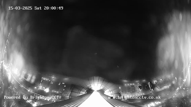 A nighttime webcam view showing a bright, illuminated street or pathway leading into a distance, with faint outlines of structures on either side. The image is predominantly black and white, featuring varying levels of brightness and some blurred areas, possibly due to atmospheric conditions. A timestamp is displayed at the top, indicating the date and time as March 15, 2025, at 20:00:49. The bottom left corner includes a watermark indicating it is powered by Brighton CCTV.