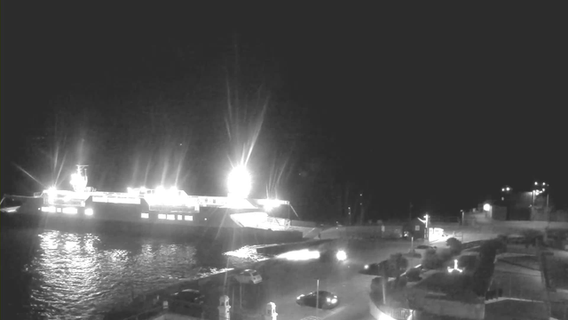A brightly lit ferry docked at a harbor at night, with reflections shimmering on the water. The scene is mostly dark, with the ferry's lights illuminating the area around it. There are several parked cars visible on the land near the dock, and faint outlines of structures, possibly buildings or a wall, are seen in the foreground and background. The overall atmosphere conveys a quiet nighttime setting by the waterfront.