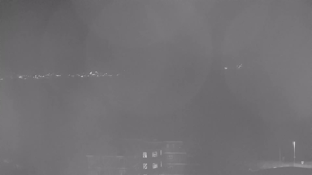 A nighttime webcam view shows a dark scene with faint lights in the distance suggesting a city skyline. In the foreground, a low, dark structure partially obstructs the view, resembling a building or complex. The overall image has a monochromatic appearance, emphasizing the limited visibility and soft glow of the distant lights against the dark sky.