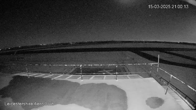 A dark image, taken from a webcam at the Leicestershire Aero Club, depicts a night scene. In the foreground, there is a flat, dark surface with a slight texture, possibly a landing area or runway. A low fence runs along the edge of the surface, and there are several white lines indicating parking spaces. In the background, there is a wide, empty field with a few faint lights visible in the distance against a mostly dark sky, which has some stars. The date and time displayed in the corner indicate it is March 15, 2025, at 21:00:13.