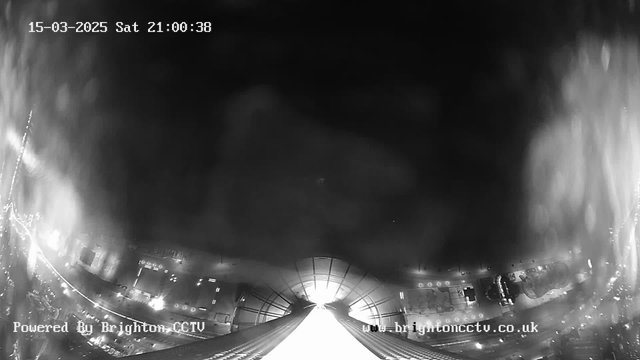 A black and white image from a webcam captures a view from a high vantage point. The scene features illuminated streets and buildings below, with a bright light source in the center suggesting an opening or passage. The upper portion of the image is darker, while light reflects off surfaces. The timestamp in the top left corner shows it is March 15, 2025, at 21:00:38. The bottom left corner has a watermark indicating it is powered by Brighton CCTV.